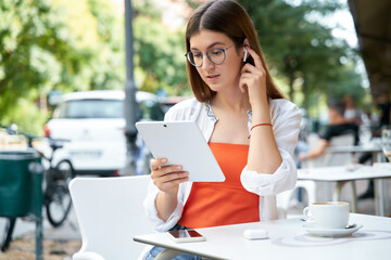 Pensive business woman in earphones using digital tablet watching training courses sitting in cafe. Successful freelancer having video call working online at workplace. Modern technology concept 