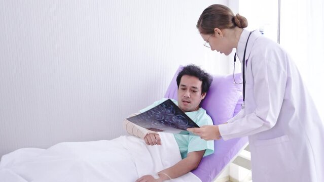 Caucasian female doctor explaining brain X-ray film to Asian male patient listening in hospital bed. Care by expert doctors. Diagnosis of neurodegenerative diseases a blood vessel in brain or cancer