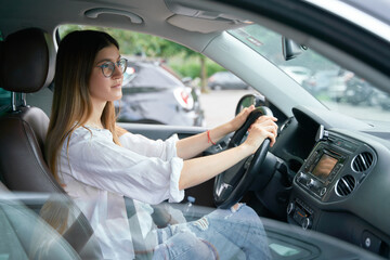 Fototapeta na wymiar Side view of the beautiful young happy smiling woman driving her car and looking at the road. Portrait of the positive woman wearing glasses keeping steering wheel in the car