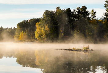 Fototapeta na wymiar A foggy and peaceful morning on a small lake in Maine during fall foliage in New England. The colorful trees with the sunshine over the pond is reflective and beautiful. 