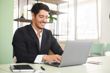 Optimistic ethnic man in formal business wear using laptop sitting on the workplace in modern open...