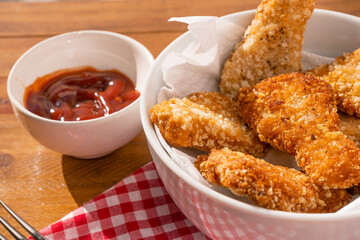 Close-up of some fried chicken nuggets in a white bowl accompanied by ketchup sauce on a wooden table. Fast food concept.