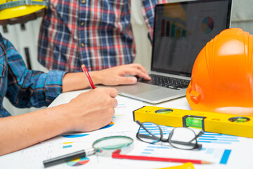 Asian engineer and partner team learn and talk with house plan project blueprint, graph and yellow helmet and engineering construction tools on table work.