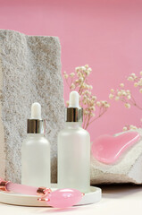 White bottles with pipettes with serum or cosmetic oil, a gouache scraper and a facial massage roller on a round pedestal against the background of natural stones and gypsophila