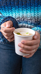 Close-up of Famele in blue sweater eating a Chicken broth soup in takeaway cup.