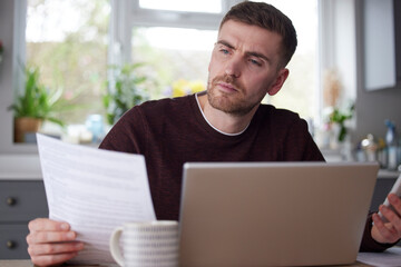 Man In Kitchen At Home Looking At Domestic Bills  With Laptop And Calculator
