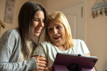 Daughter showing her mother something on the tablet and the mother is surprised while they are happily sitting on the sofa at home.