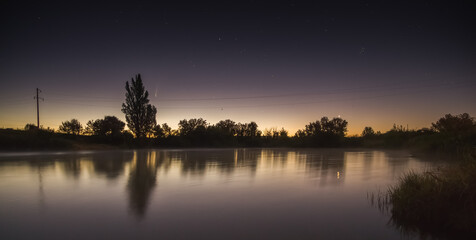 The beginning of dawn on the lake on a starry night in a clear sky with stars and comet Neowice, long exposure