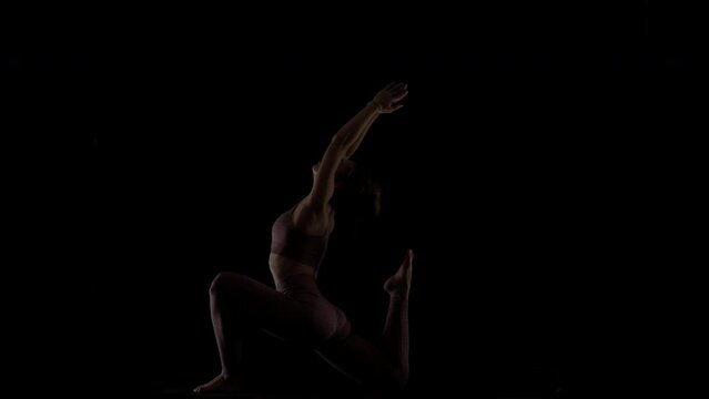 Fit woman practicing yoga poses. Silhouette girl doing exercise indoors against black background. No stress inner balance concept..