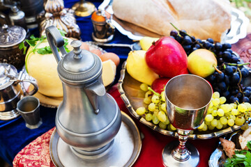 A metal jug and a glass for wine on the background of a table with fresh fruits. Food and utensils in the medieval style. Reconstruction of the life of the ancient peoples of the times of chivalry.