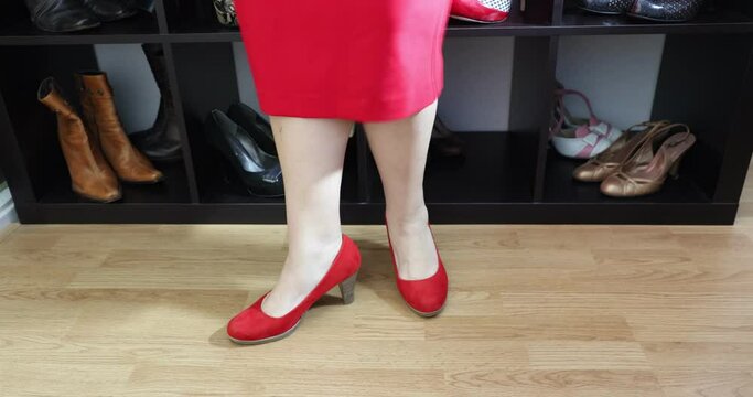 Female legs and feet of standing adult woman trying on red high heels, home dressing room with cabinet with shoes, red dress, high perspective side view. Beauty, glamour and fashion concept