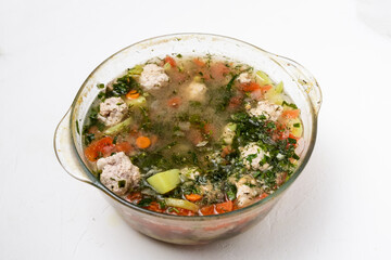 Vegetable soup with meatballs in a glass saucepan on a white background.