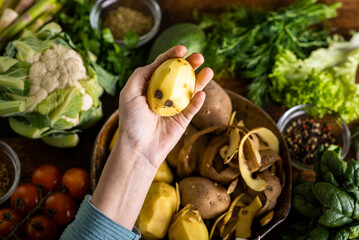 A woman holds a raw peeled potato in her hands. Various fresh vegetables lie on the table. Cooking vegetarian food.