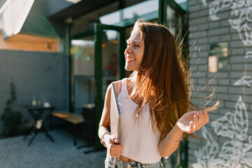 Close-up of pretty young caucasian woman looking away standing on street with laptop. Girl with brown long hair wears t-shirt. Lifestyle concept