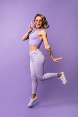 Fototapeta na wymiar Joyful european young woman showing tongue, winking and showing victory hand in studio. Blonde girl wears glasses, top and pants and bounces on purple background. Lifestyle concept