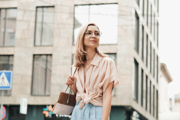 Fototapeta premium Young Elegant Woman in Glasses Walking in the City, Stylish Blonde Girl Outdoors Standing on the Street Looking Away.