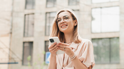 Happy Elegant Woman in Eyeglasses Holding Smartphone in Hands and Smiling, Stylish Blonde Girl with Glasses Standing Outdoors on the Street.