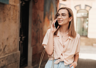 Cheerful Elegant Woman in Eyeglasses Using Smartphone and Smiling, Stylish Blonde Girl with Glasses Standing Outdoors on the Street Having Conversation Through Mobile Phone.