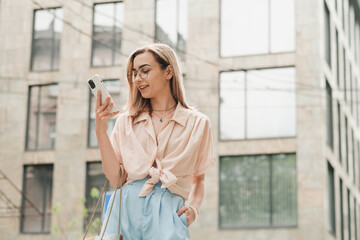 Fototapeta premium Cheerful Elegant Woman in Glasses Holding Smartphone in Hand and Smiling, Stylish Blonde Girl Outdoors Standing on the Street Looking on Cell Phone.