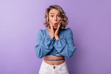 Surprised young caucasian woman looking dazedly at camera holding her hands near face of in studio. Blonde woman with wavy hair wears blouse. Perplexity concept