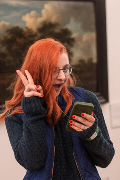 A picture of a red haired woman with a blue sweater and a phone standing in a museum in front of a painting 