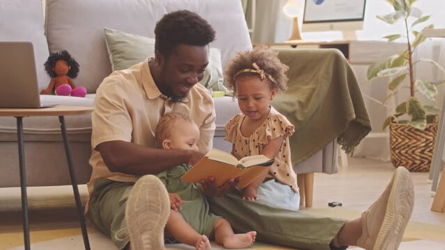 African American man reading book to his toddler son and daughter sitting together on rug in cozy living room