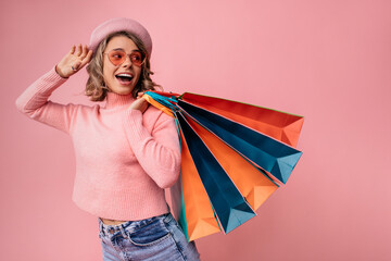 Beautiful young caucasian blonde woman standing over pink background holding shopping bags. Blonde wears sunglasses, beret, sweater and jeans. Purchase concept