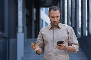 Frustrated and upset african american man outside office building trying to make a purchase in an online store, man using smartphone and bank credit card, to transfer money and buy online.