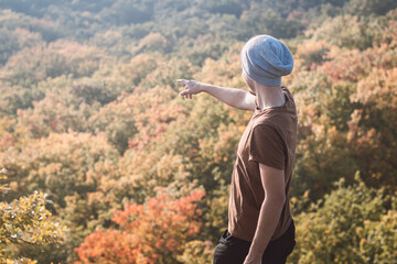 Attractive youthful man in a brown T-shirt and gray cap stands on a rock and points to the leafy trees playing with all the colors of autumn. Wild Sarka, Prague, Czech Republic