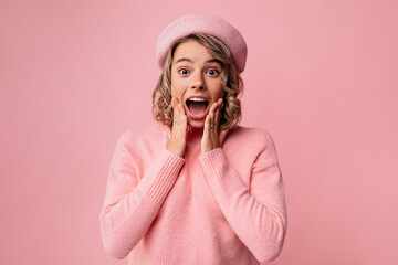 Nice young caucasian lady holding hands on cheeks with mouth open is surprised on pink background. Blonde woman wavy hair wears beret and sweater. Shock concept