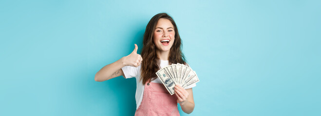 Smiling happy woman holding money, dollar bills and showing thumb up, recommending fast cash loan...