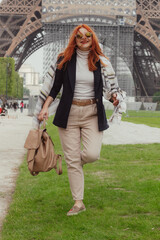 A picture of a smiling, red haired woman, walking with a brown bag in her hand and wearing a blue...