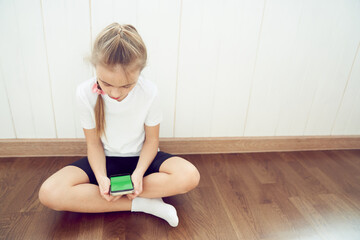 A girl who sits on the floor and uses a phone with a green screen. The child is sitting and looking into a smartphone with a colored key. High quality photo