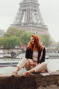 A picture of a red haired woman sitting on a stone bridge across the Eiffel Tower, while looking away from the camera and wearing a blue blazer, white t-shirt and cream pants 