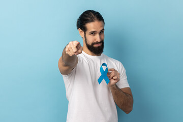 Portrait of responsible handsome man with beard wearing white T-shirt holding blue awareness, disease symbol, pointing to camera, support. Indoor studio shot isolated on blue background.