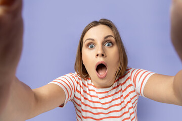 Portrait of shocked woman in striped T-shirt taking selfie picture, point of view, looking at the camera with open mouth, making front selfportrait. Indoor studio shot isolated on purple background.