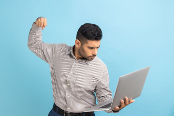 Angry businessman punching laptop screen, looking with mad expression, boxing threatening to hit while having online conversation, wearing striped shirt. Indoor studio shot isolated on blue background