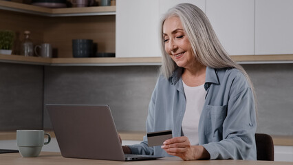 Happy old female caucasian consumer grey-haired granny shopper pay internet order on laptop sits in kitchen hold banking card use online banking app donating money deposit rejoice approved credit loan