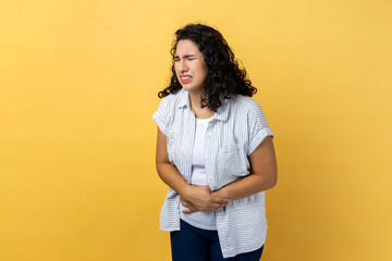 Portrait of unhealthy sick beautiful woman with dark wavy hair standing, holding her belly with hands, stomach cramps or period pain. Indoor studio shot isolated on yellow background. - obrazy, fototapety, plakaty