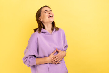 Obraz premium Portrait of happy woman with dark hair holding belly, laughing, hearing funny joke or anecdote, being in good mood, wearing purple hoodie. Indoor studio shot isolated on yellow background.