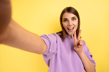 Beautiful smiling woman taking selfie, looking at camera POV, point of view of photo, showing victory gesture, wearing purple hoodie. Indoor studio shot isolated on yellow background.