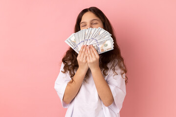 Portrait of rich satisfied little girl wearing white T-shirt holding big fan of money, smelling dollar banknotes with pleasure, keeps eyes closed. Indoor studio shot isolated on pink background.