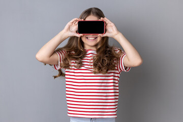 Portrait of little girl wearing striped T-shirt covering eyes with cellphone and smiling, unknown child hiding face with mobile phone, anonymous user. Indoor studio shot isolated on gray background.
