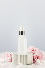 Glass dropper bottle with serum on marble podium with tender pink roses and rose petals. Natural beauty product based on rose flowers, fermented cosmetic. Soft focus style, copy space, vertical format