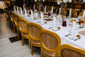 A large long table covered with a white tablecloth with dishes, forks, spoons, knives, food stands inside the restaurant near the chairs. Restaurant photo.
