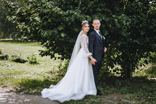 A stylish groom in a blue plaid suit and a beautiful bride in a white lace dress with a diadem, a bouquet in her hand gently hug in a park in nature. Wedding photography, portrait of smiling newlyweds