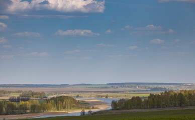 Sunlight through the clouds illuminates the young greenery of bushes and trees. Rural spring landscape with a river, clouds over the horizon.