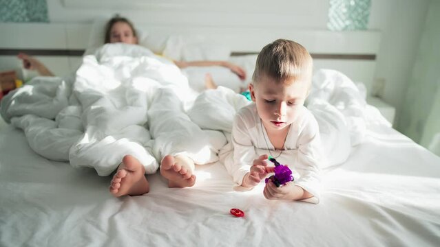 Home life brother and sister or mother in bedroom lie on bed with white linens. Sun rays in home interior in morning. Baby boy is playing with transformer robot toy. Family happy leisure activity.