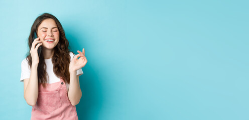 Fototapeta na wymiar Portrait of cute and stylish girl laughing while chatting on phone, holding smartphone near ear and smiling happy, having casual conversation, standing over blue background