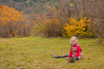 a little preschooler in a sundress and a red turtleneck, pleased with a smile, sits on the grass on a warm autumn day against the background of trees with yellow foliage